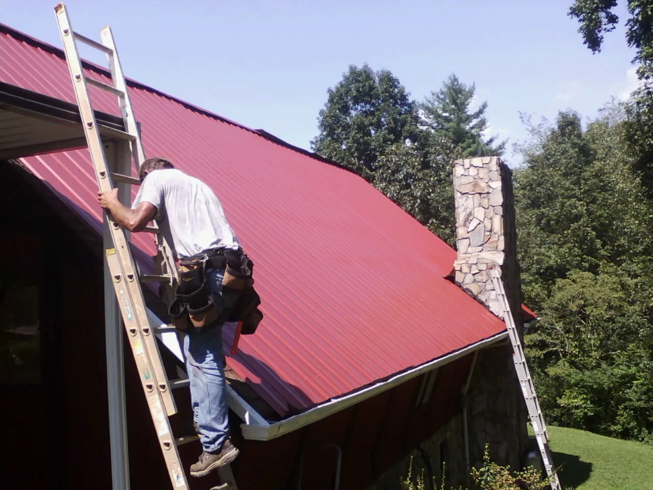 Roofer on a ladder beside a red metal roof and stone chimney — Soffit & Fascia Repair by Roofing Brothers in Glenfield